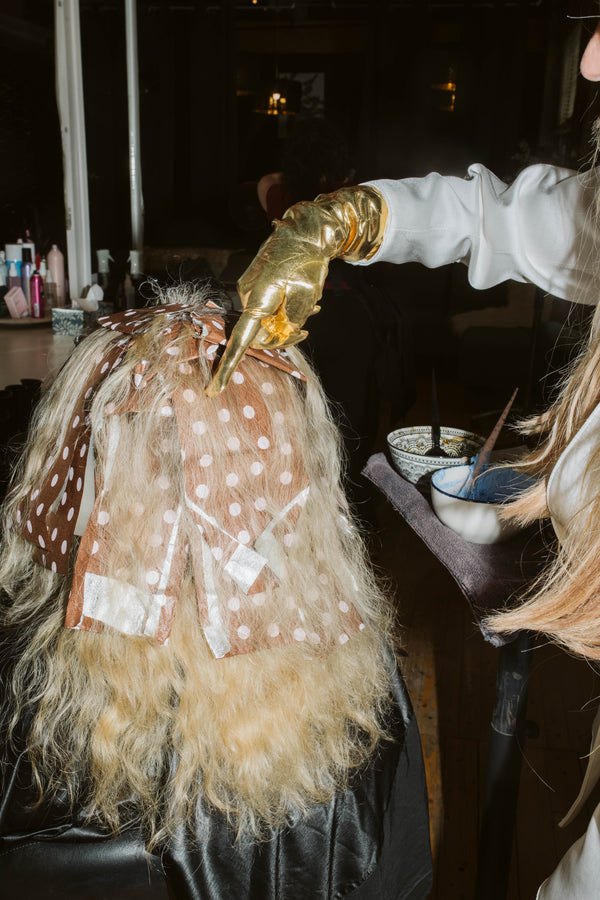 A high-contrast flash photo of a gold-gloved hand pointing at a spot on a client's foiled blonde hair.