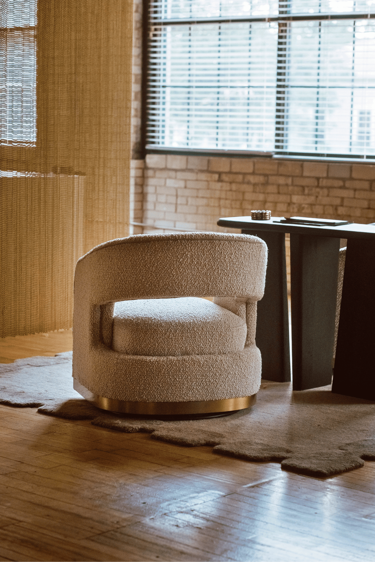 A modern bouclé accent chair in a warm cream color with a gold metal base, positioned on a rustic wood floor in a stylishly decorated room with a brick wall and window blinds.