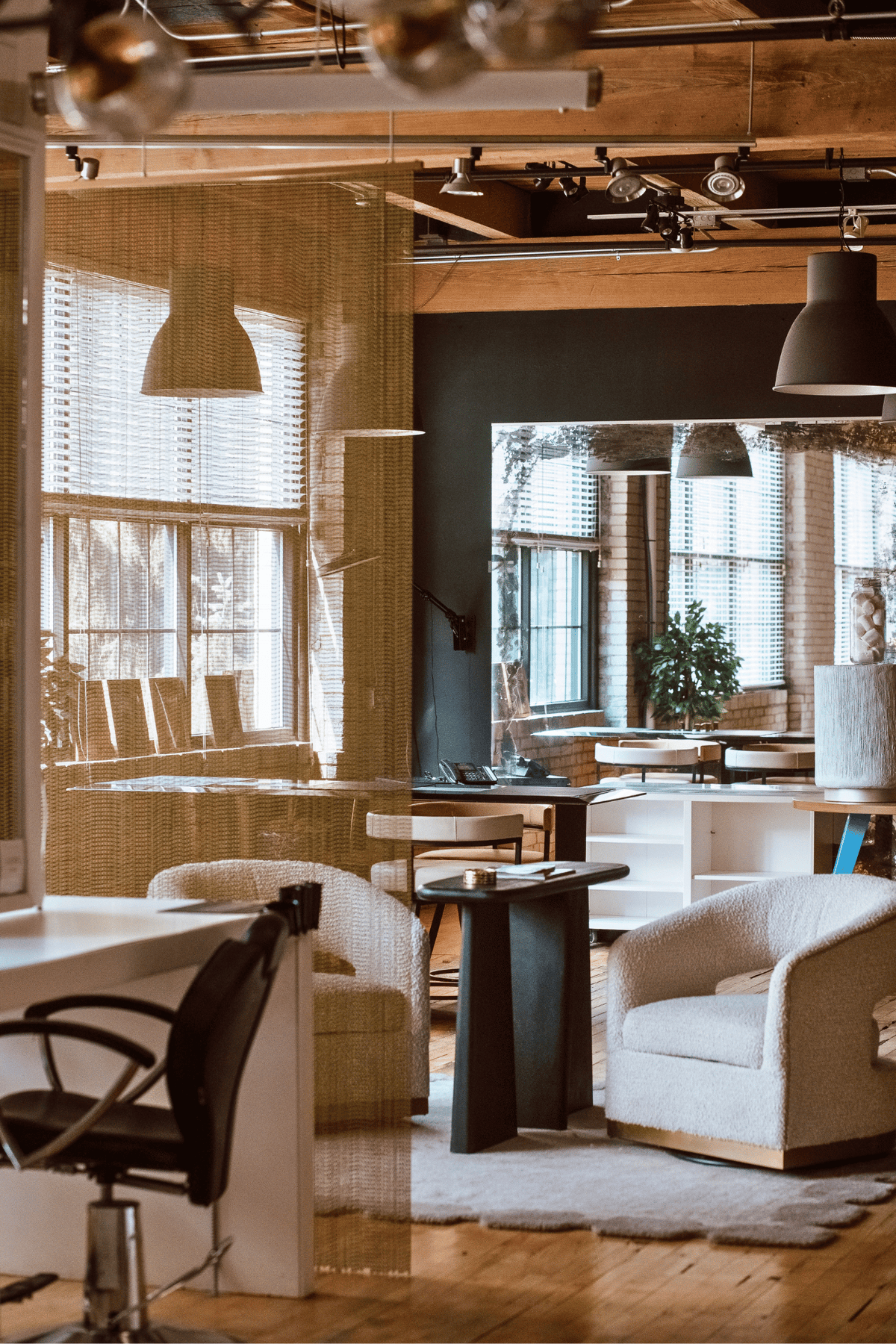 A modern interior space with exposed wood ceiling beams, pendant lights, large windows with blinds, and upholstered seating including cream armchairs, a black office chair, and a small round table.