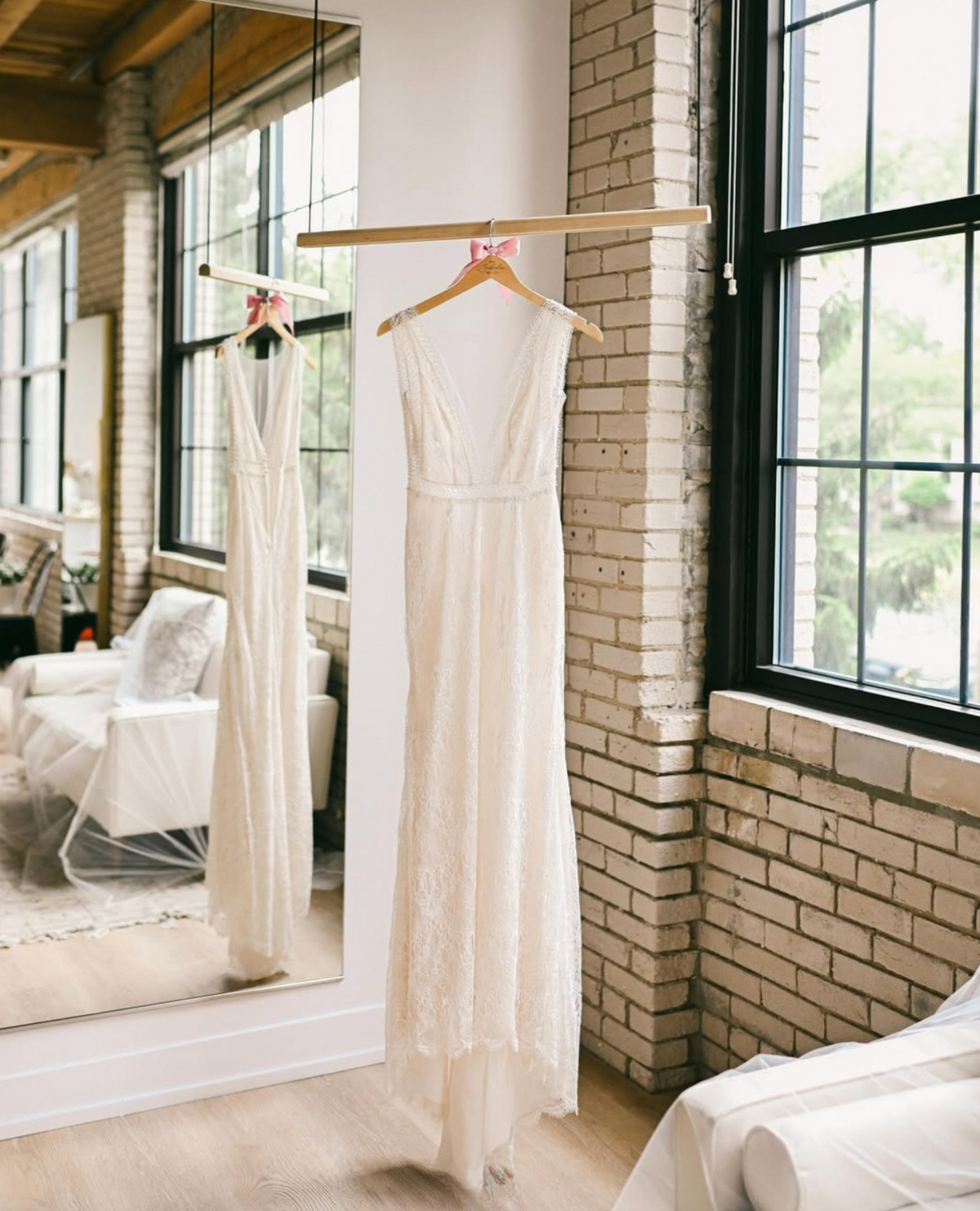 An ivory lace sleeveless gown with a deep V-neck and waistband detail hangs on a wooden hanger from a clothing rack in a bright room with a brick column and large windows. A mirror on the left reflects the dress.
Photo by @lindseymareephotography