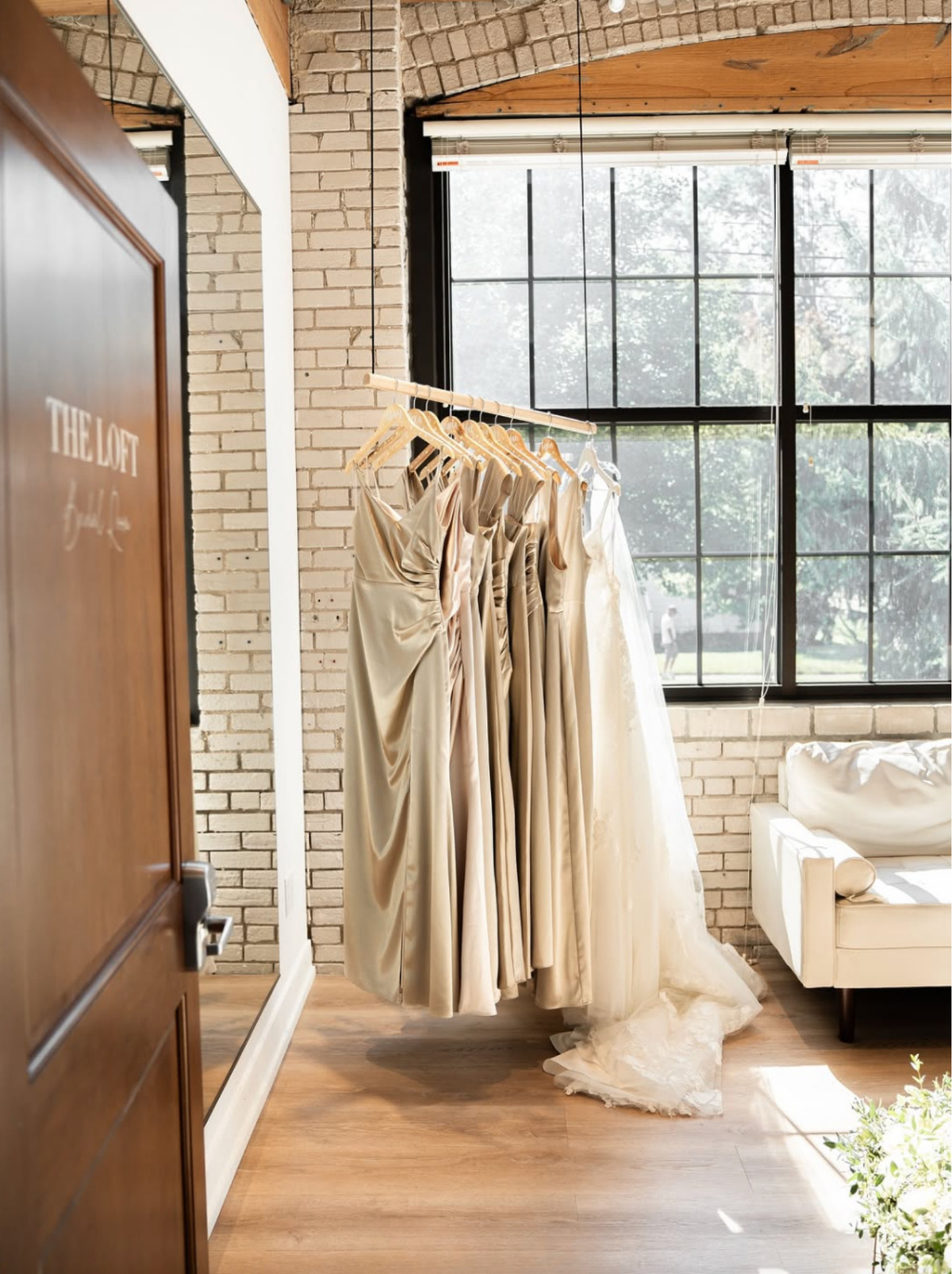 A set of champagne and ivory satin bridesmaid dresses hanging on wooden hangers from a clothing rack inside a boutique. The boutique features a white brick wall, a large black-framed window, and a wooden floor.
Photo by @windandwavesmedia_