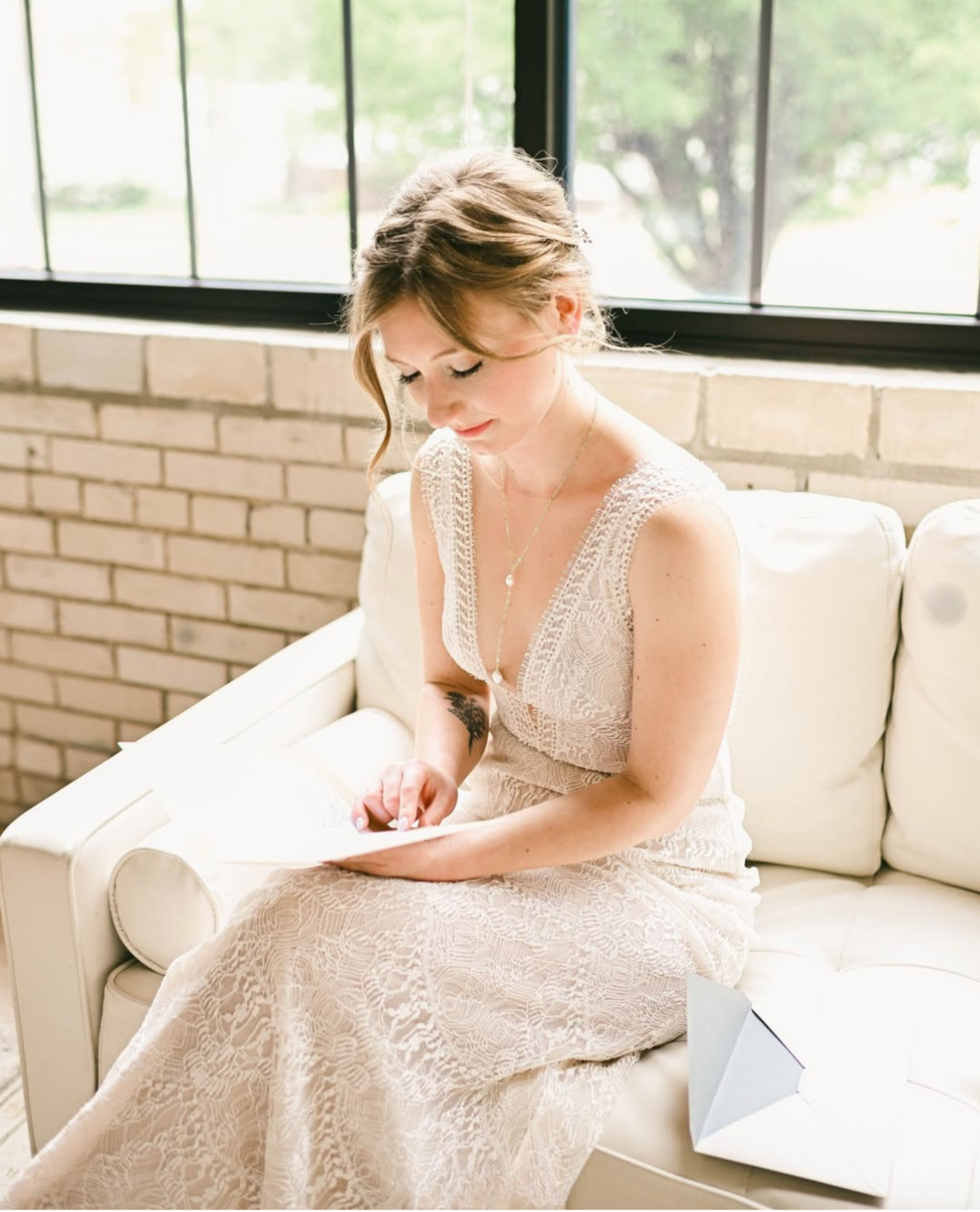 A woman in a sleeveless lace wedding dress sits on a cream sofa near a window, holding a small white envelope or card.
Photo by @lindseymareephotography