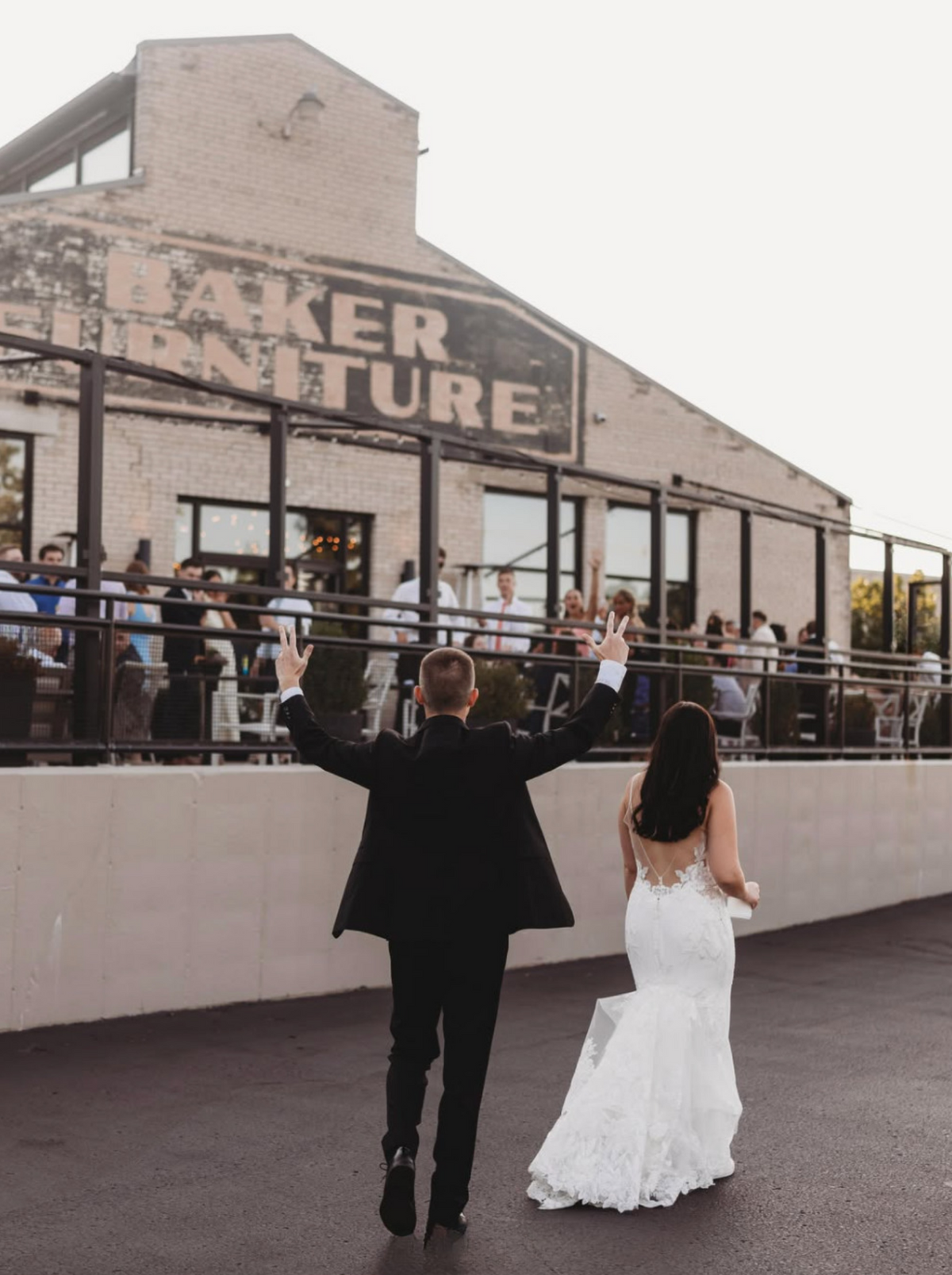 A bride in a white lace mermaid wedding gown and a groom in a black suit walk away from the camera on a rooftop terrace, with a brick building and guests in the background.
Photo by @mandmterryphotography.