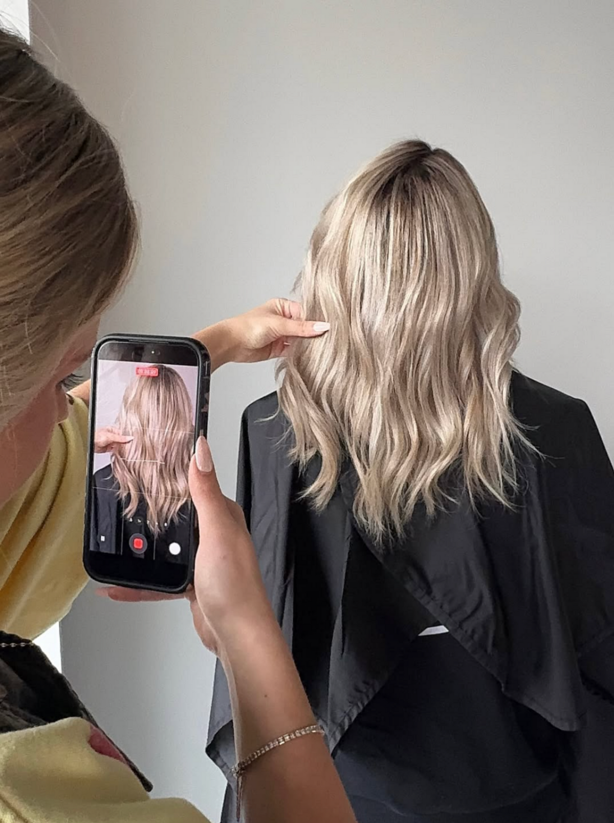 A person with shoulder-length blonde wavy hair, viewed from the back while wearing a black salon cape. Another person on the left holds up a smartphone showing a mirror-style reference photo of the hair color.
Blonding by Blythe