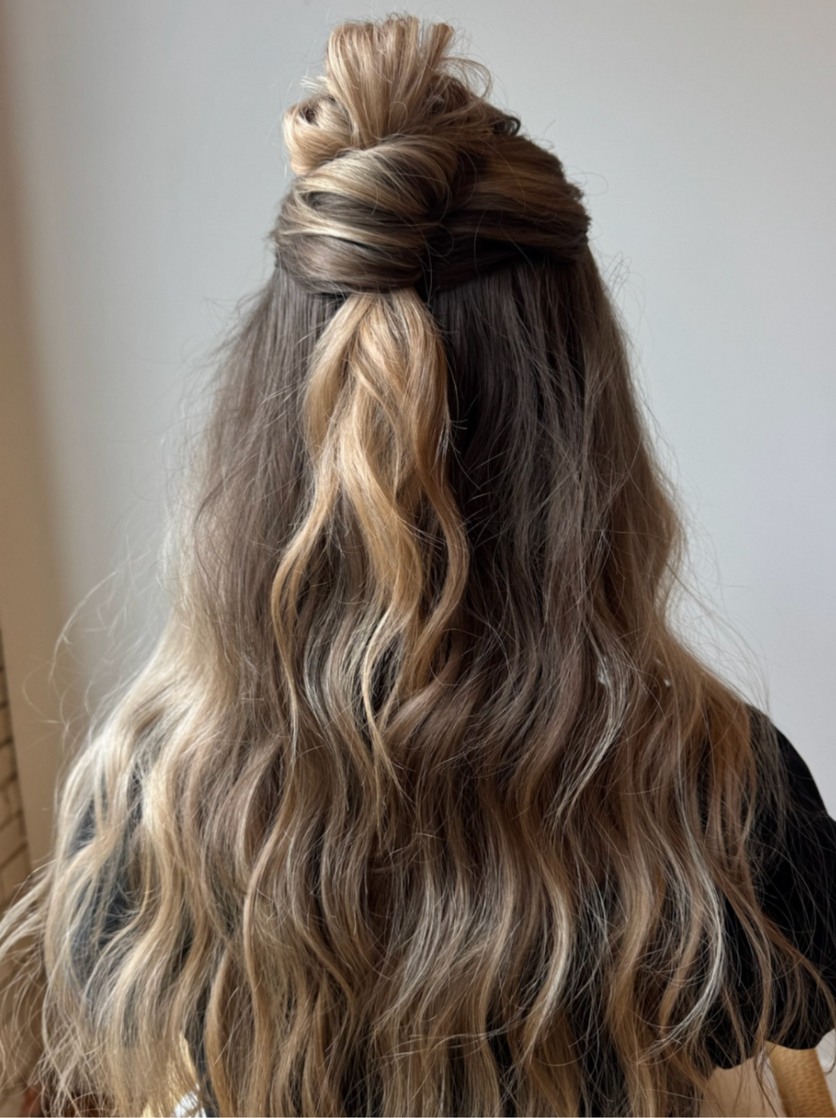 Long wavy hair with a dark brown to light blonde ombré color, styled with a half-up bun and loose curls, shown from the back.