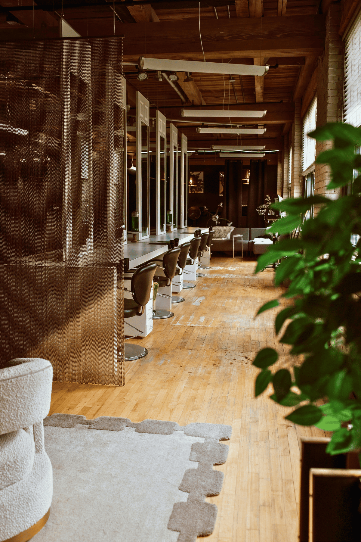 A long, narrow salon or studio interior with wooden floors, hair stations, mirrors, pendant lights, and a large green plant in the foreground. No person is visible.