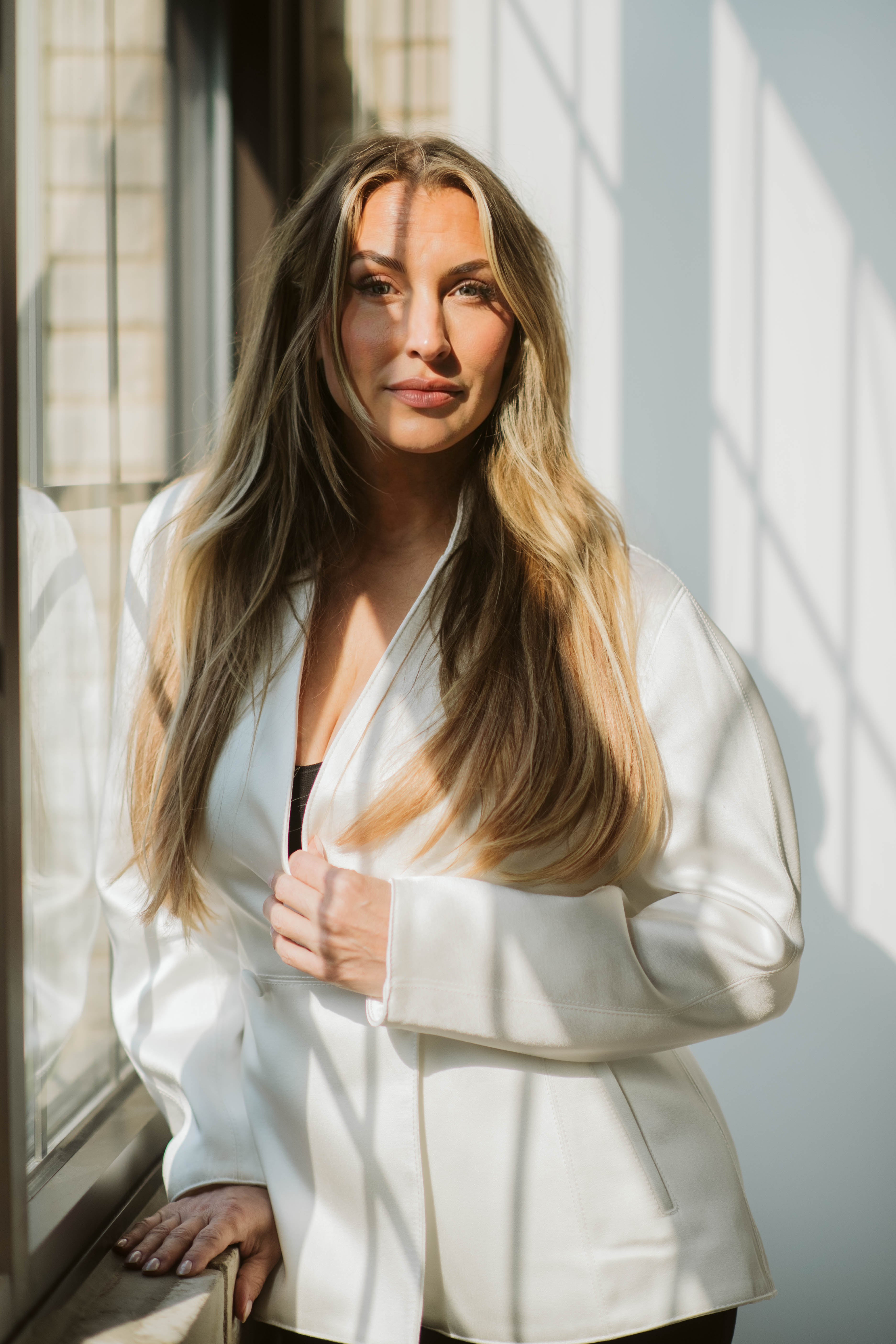 A woman with long blonde hair wearing a white satin blazer, standing by a window with warm sunlight casting shadows on the wall.