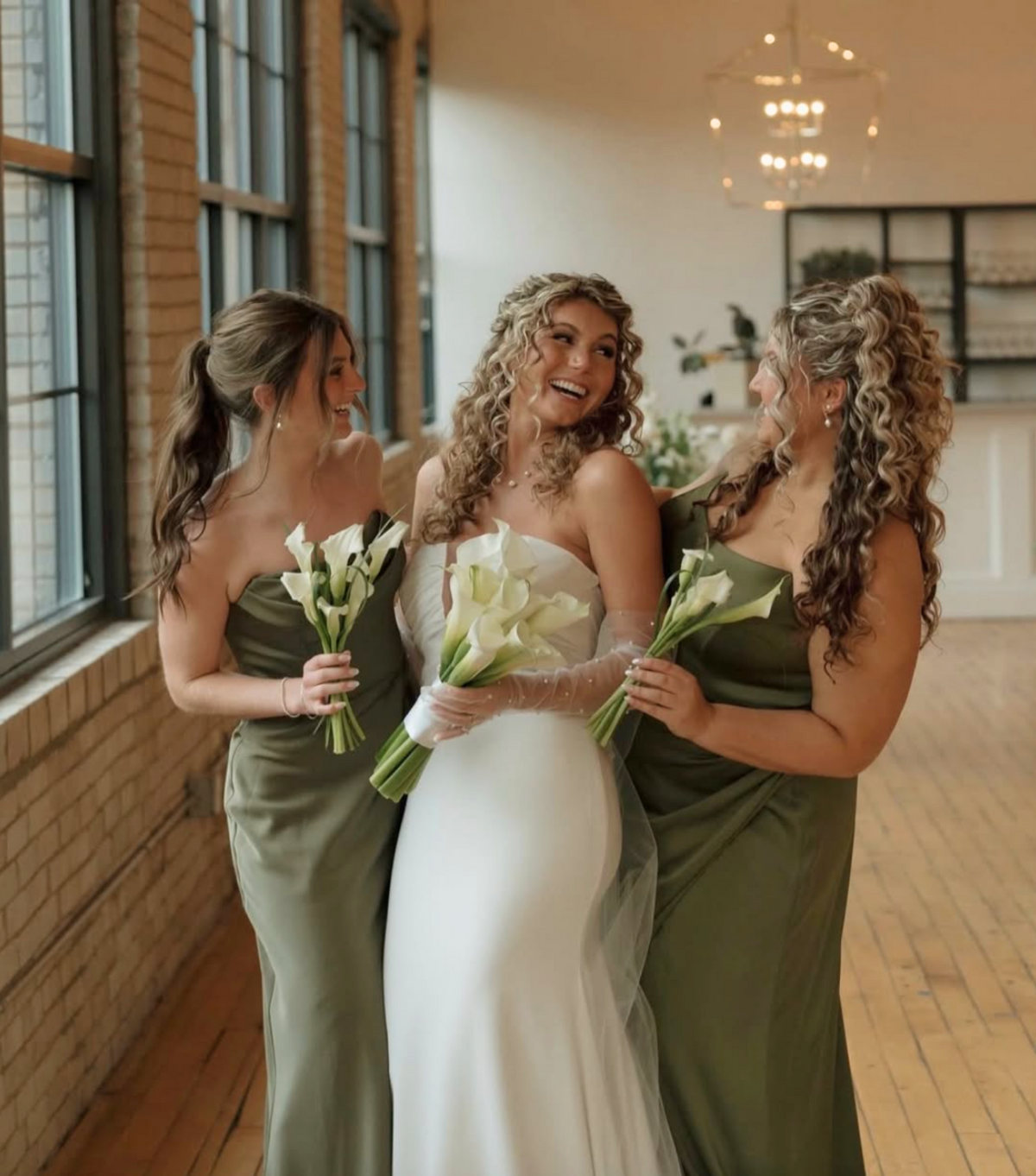 Three women posing together in a bright indoor space with brick walls and wood floors. The central woman wears a strapless white wedding gown and holds a bouquet of white calla lilies, while the two women beside her wear matching olive green strapless bridesmaid dresses and also hold bouquets of white lilies.
Photo by @imagesbyhannahgrace.