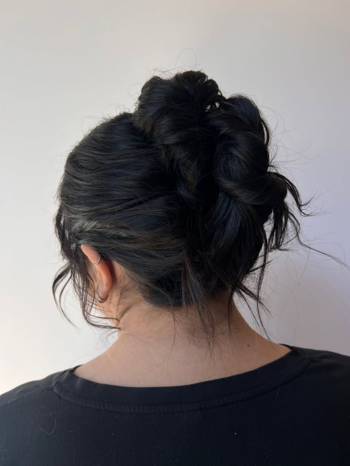 Back view of a person with long, dark, glossy hair styled in a messy bun, wearing a black t-shirt against a plain light gray wall.
