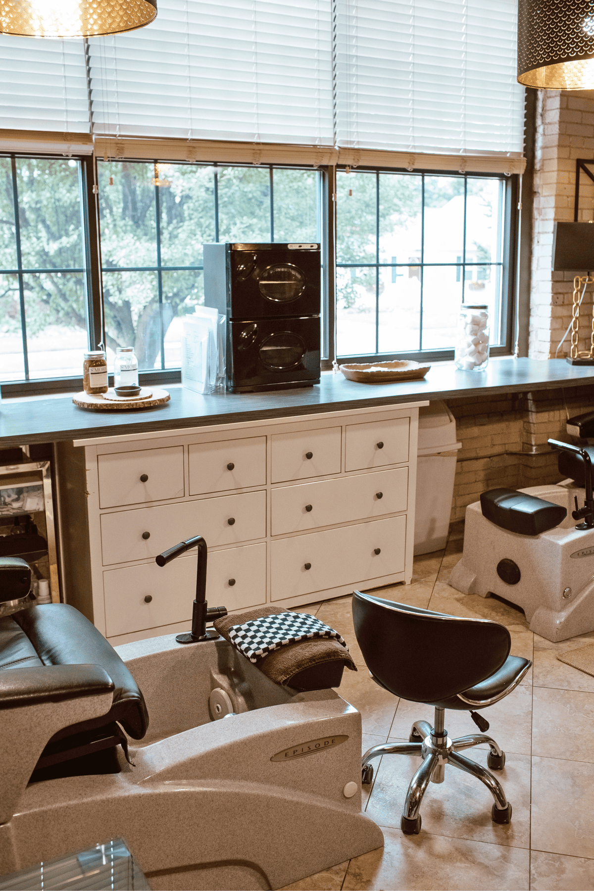 A modern nail spa or salon interior with multiple treatment chairs, a white storage cabinet, windows with blinds, and countertop tools. No single retail product is isolated, and the scene includes a real environment with furniture and fixtures.