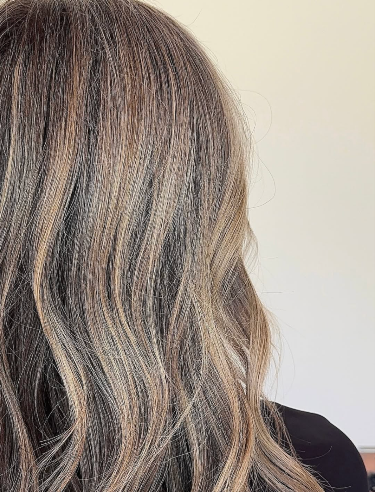 Close-up of a person's shoulder-length wavy hair with a balayage-style blend of dark brown, gray, and warm blonde highlights, wearing a black top against a plain off-white wall.