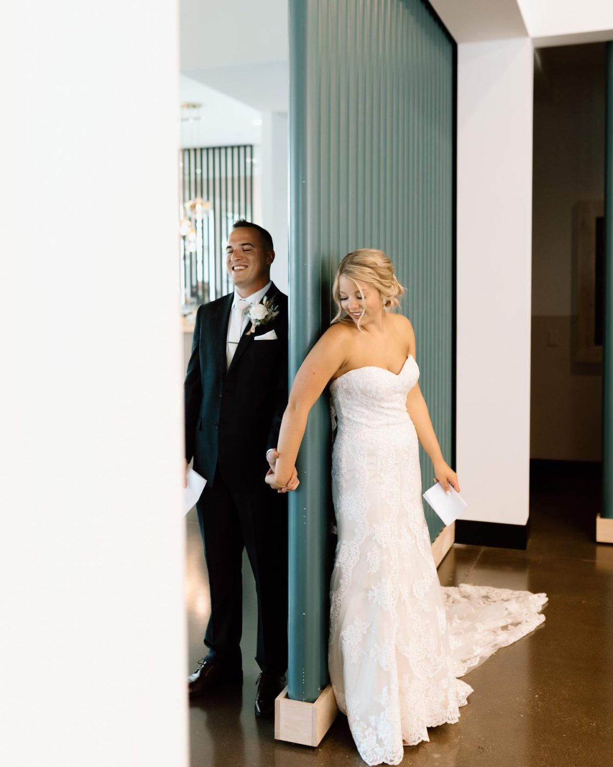 A bride in a strapless white lace wedding gown and a groom in a black tuxedo holding hands, standing in a hallway next to a teal vertical panel.