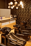 A salon wash station with multiple black leather shampoo chairs and glossy black sink bowls, set in a modern room with a patterned dark wall, wood floor, and a chandelier overhead.