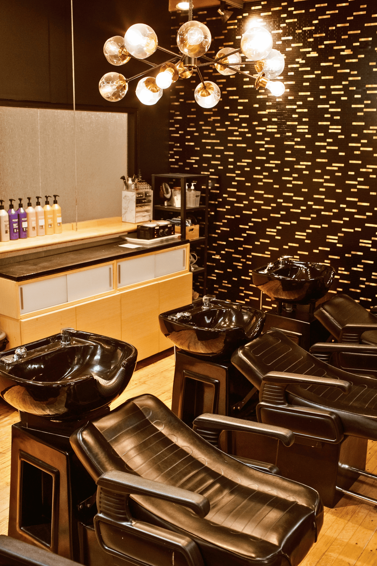 A salon wash station with multiple black leather shampoo chairs and glossy black sink bowls, set in a modern room with a patterned dark wall, wood floor, and a chandelier overhead.