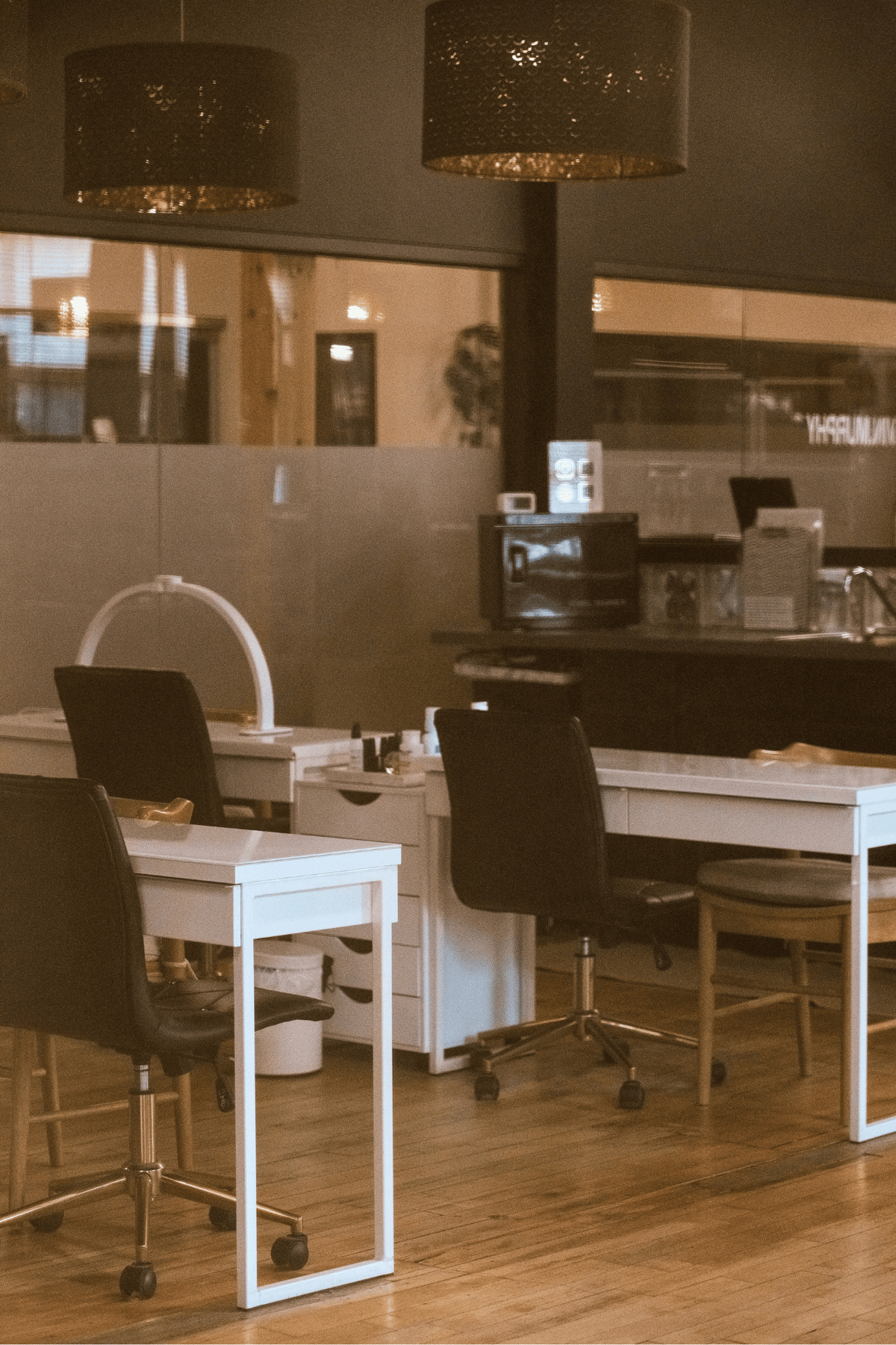 A modern nail salon interior with white manicure stations, black office chairs, wood flooring, and hanging pendant lights. Nail bachelorette parties in Holland, MI.